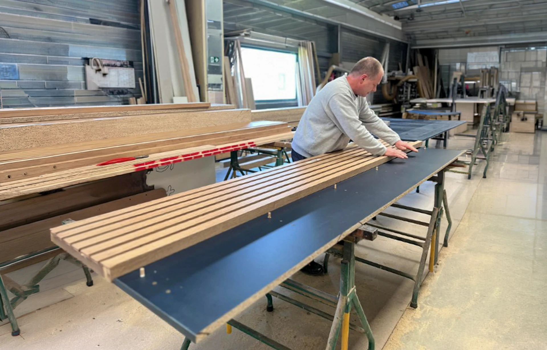 Atelier menuiserie intérieur avec artisan en salopette travaillant sur ordinateur portable, prenant notes avec crayon sur plans techniques